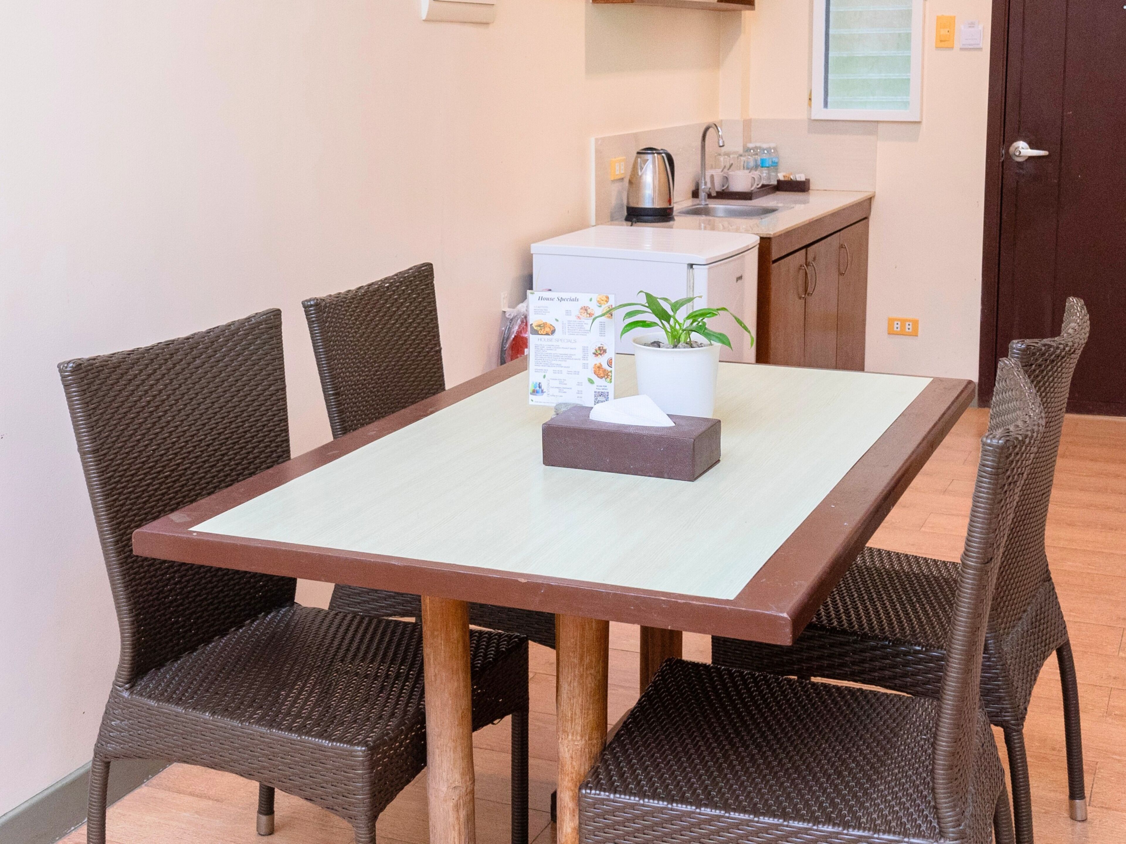 Un comedor con una mesa, cuatro sillas, una planta en maceta y una cocina al fondo.