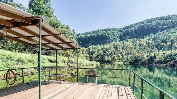 Una terraza de madera con barandilla metálica que da a un lago tranquilo, rodeada de abundante vegetación y colinas.