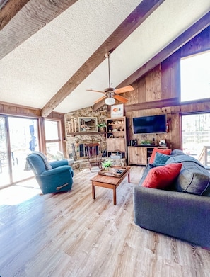 A spacious living room with wooden floors, a ceiling fan, a stone wall, and a television.