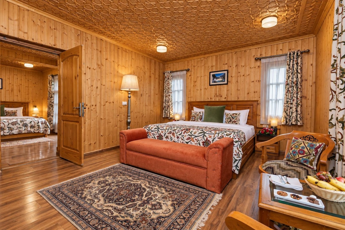 Quarto com painéis de madeira, uma cama, um banco, uma cadeira e uma mesa com cesta de frutas.