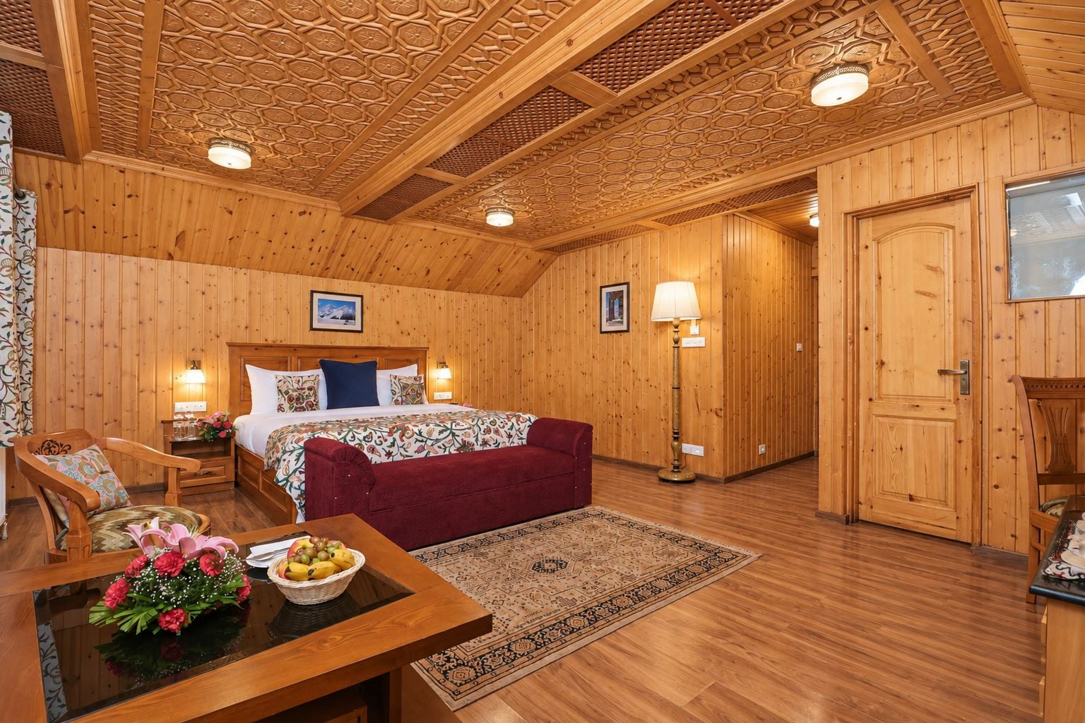 Una habitación de madera con una cama, un sofá rojo, una mesa con flores y una alfombra estampada.