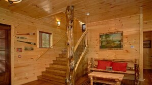 A wooden cabin interior with a staircase, a bench with red cushions, and a painting on the wall.