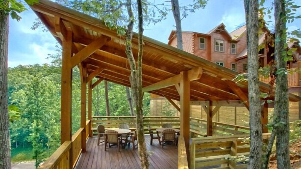 A wooden deck with outdoor seating, surrounded by trees and a building in the background.