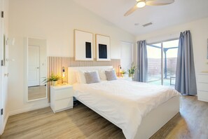 Une chambre à coucher avec un grand lit, des tables de chevet, un ventilateur de plafond et une vue sur l’extérieur, accessible par une porte-fenêtre coulissante.