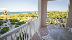 A balcony with a glass table and chairs, overlooking a beach and lush greenery.
