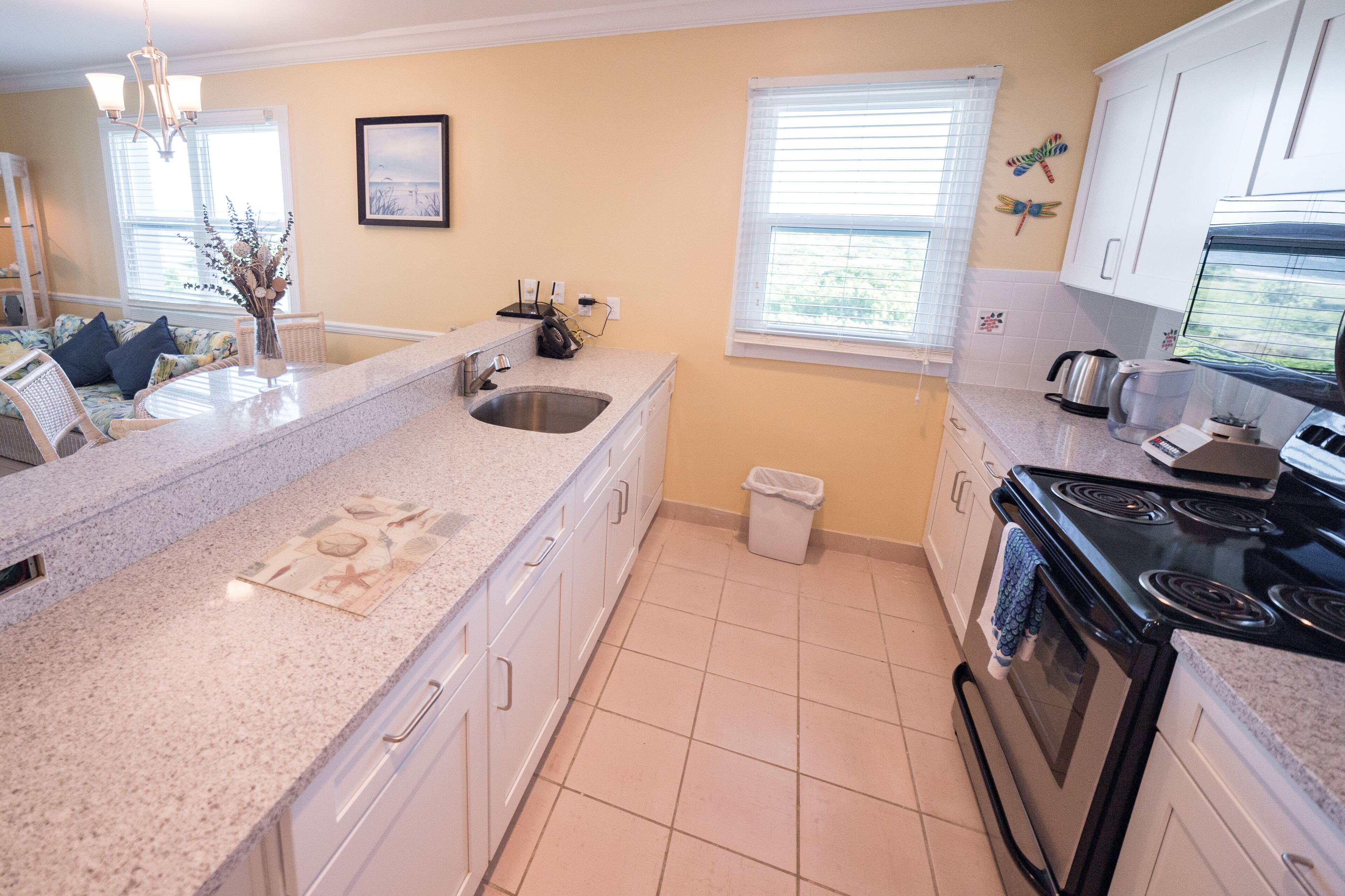 A kitchen with a granite countertop, stainless steel sink, white cabinets, a black stove, and a window with blinds.