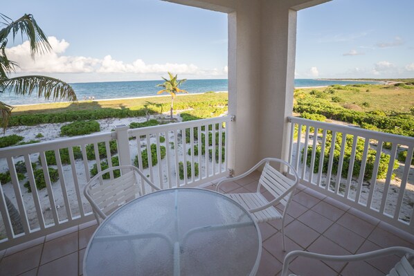 A balcony with a glass table and white chairs, overlooking a beach and ocean. - 2 Bdrm Condo Located At Northwest Point Resort. Enjoy Beach-Pool -Hot Tub-views (Wheeland Settlement)
