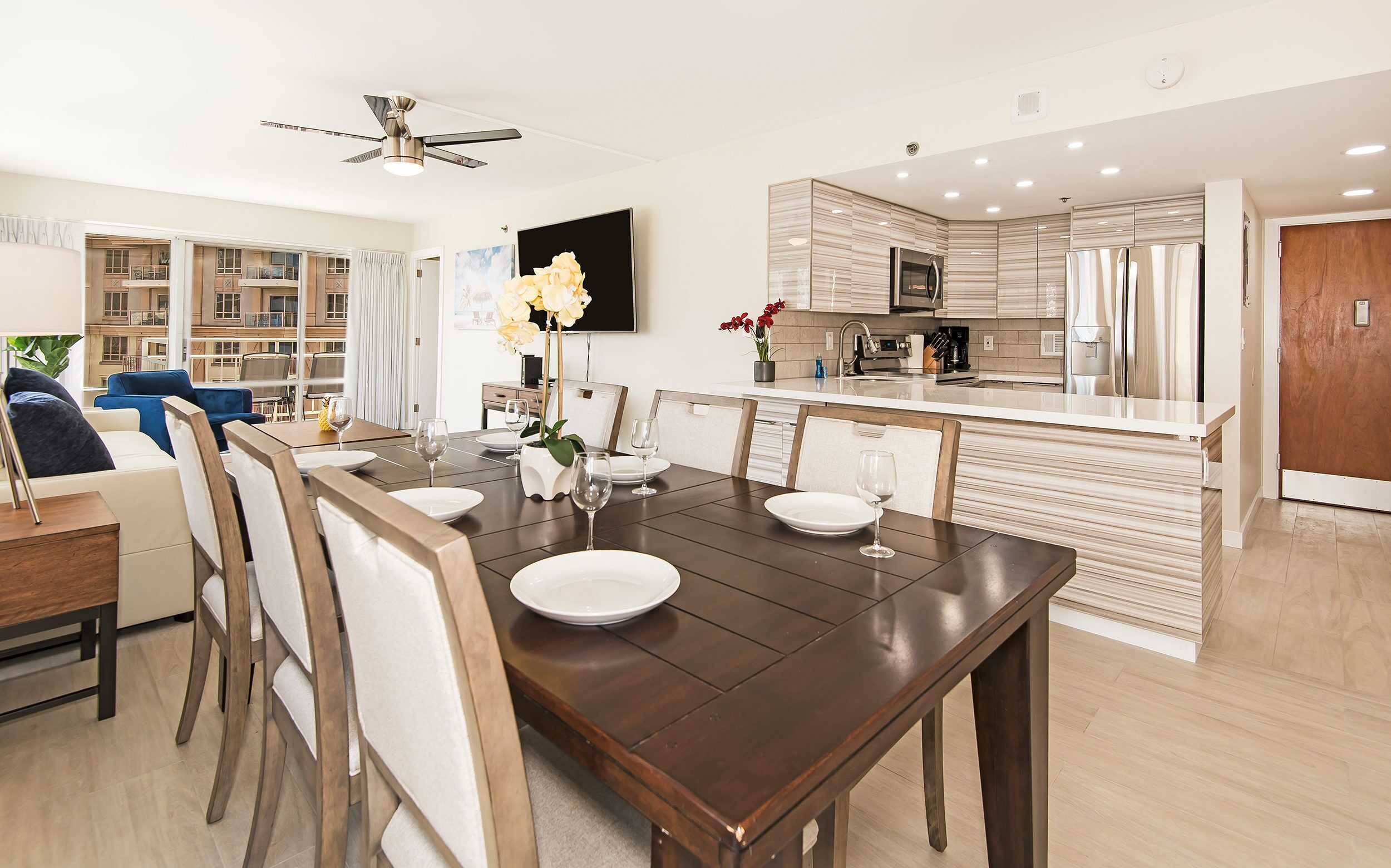 A modern dining area with a wooden table set for four, a kitchen with a built-in oven and refrigerator, and a living area with a sofa and a TV.