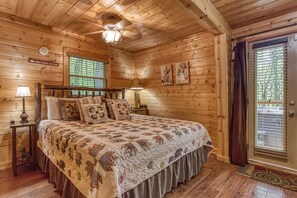 Quarto de cabana de madeira com uma cama, mesa de cabeceira, luminária, ventilador de teto e janela com persianas.