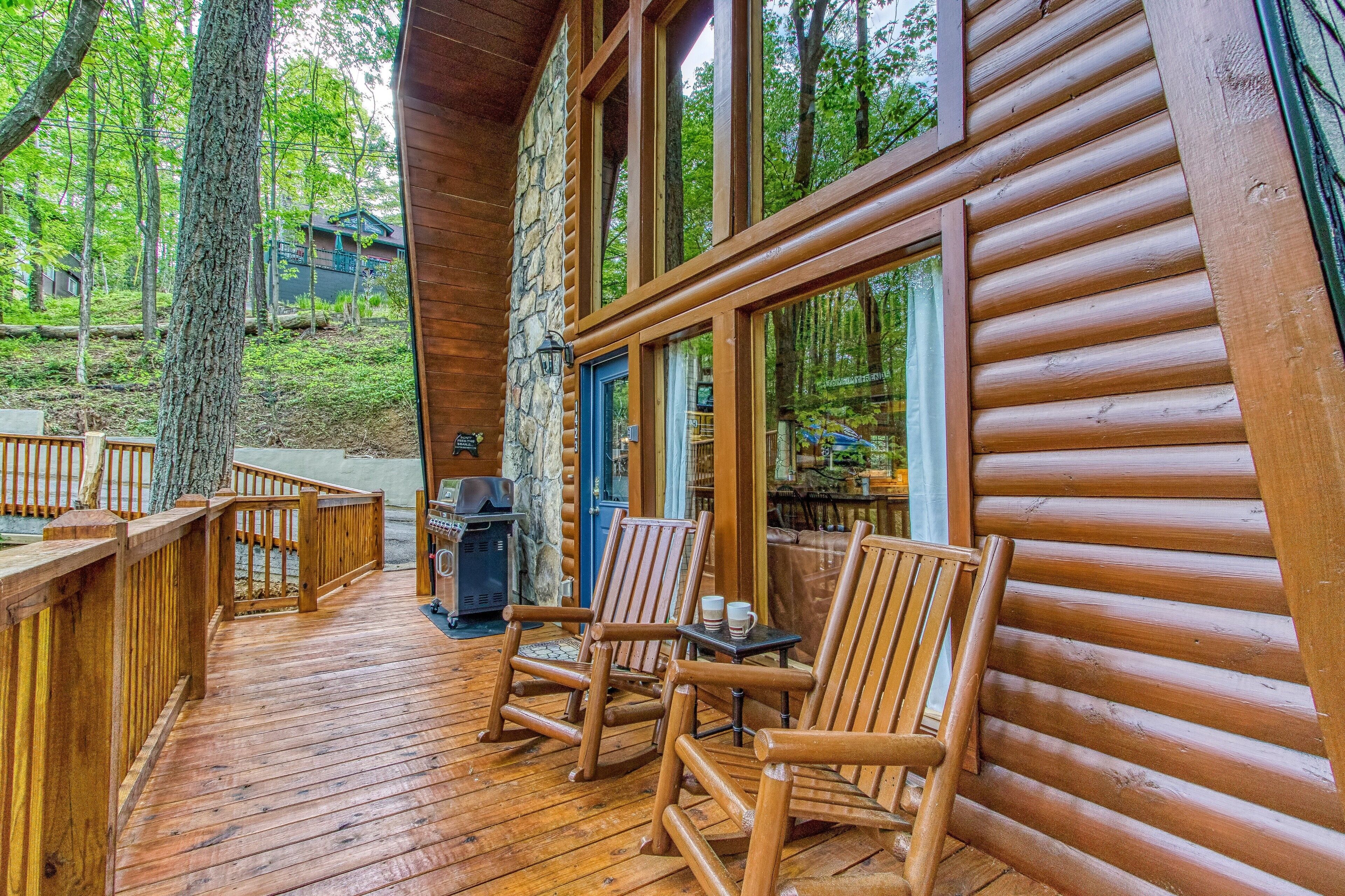A wooden deck with outdoor seating, a stone wall, and large windows overlooking a forested area.