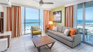 A modern living room with a sofa, armchair, and coffee table, large windows with curtains, a ceiling fan, and a view of the beach.