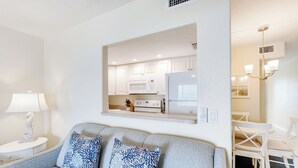 A living room with a grey sofa, two patterned cushions, a white side table with a lamp, and a view into a kitchen with white cabinets and appliances. - OCEAN'S REACH 2B4: 1-Bdrm, Top Floor w/Gorgeous Views, All Amenities Open! (Sanibel Island)