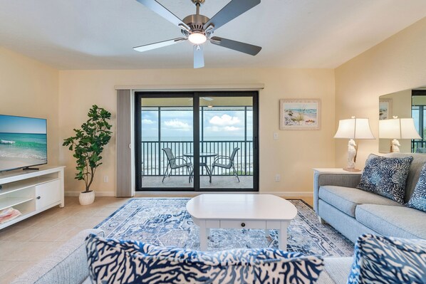 A living room with a ceiling fan, a TV, a potted plant, a balcony with chairs, a sofa, and a patterned rug.