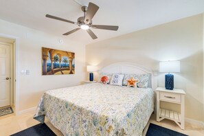 A bedroom with a bed, a ceiling fan, a nightstand, a lamp, and a painting on the wall.