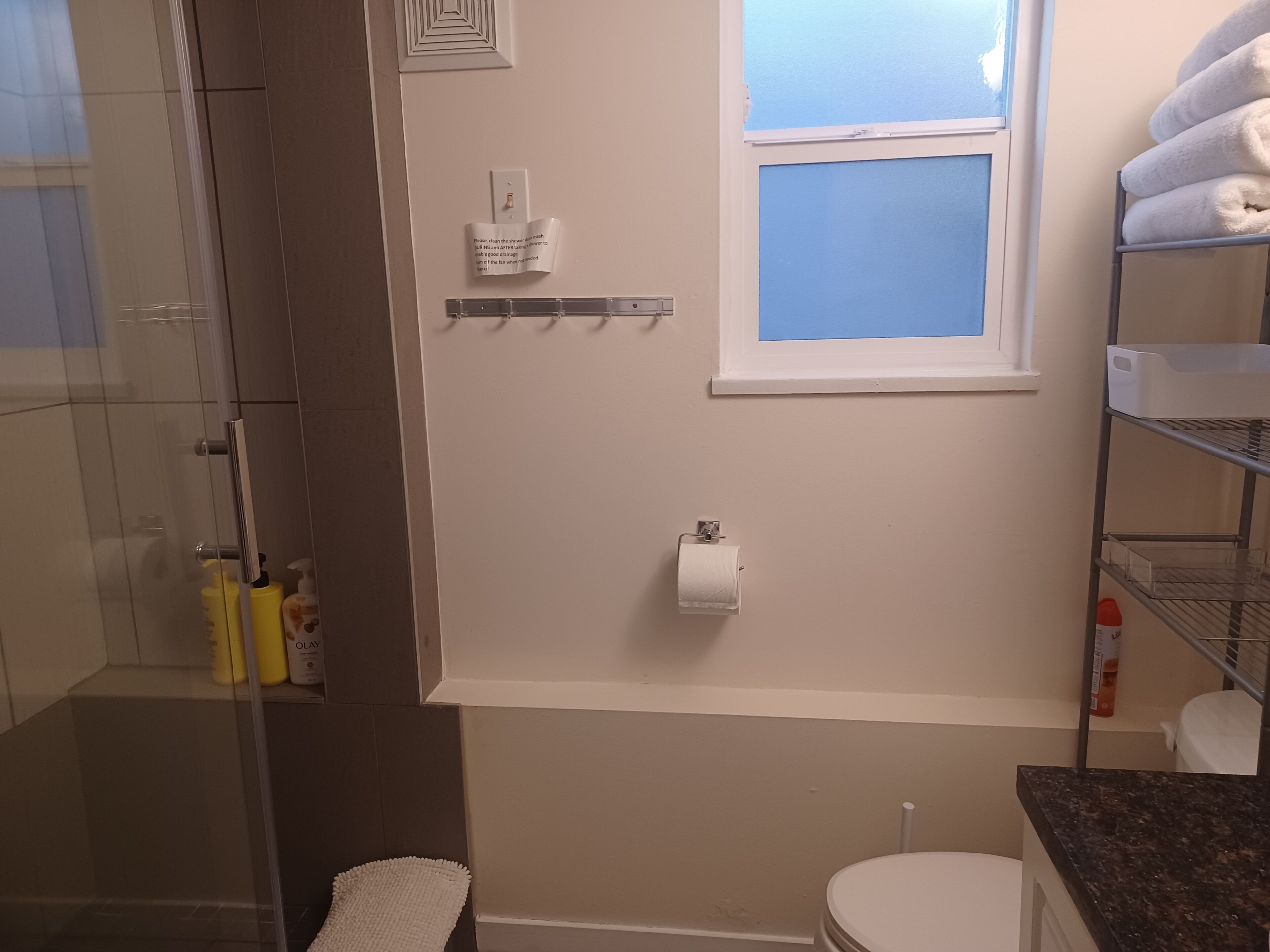 A bathroom with a glass shower enclosure, a toilet, a sink with a mirror, a window, and shelves with towels.