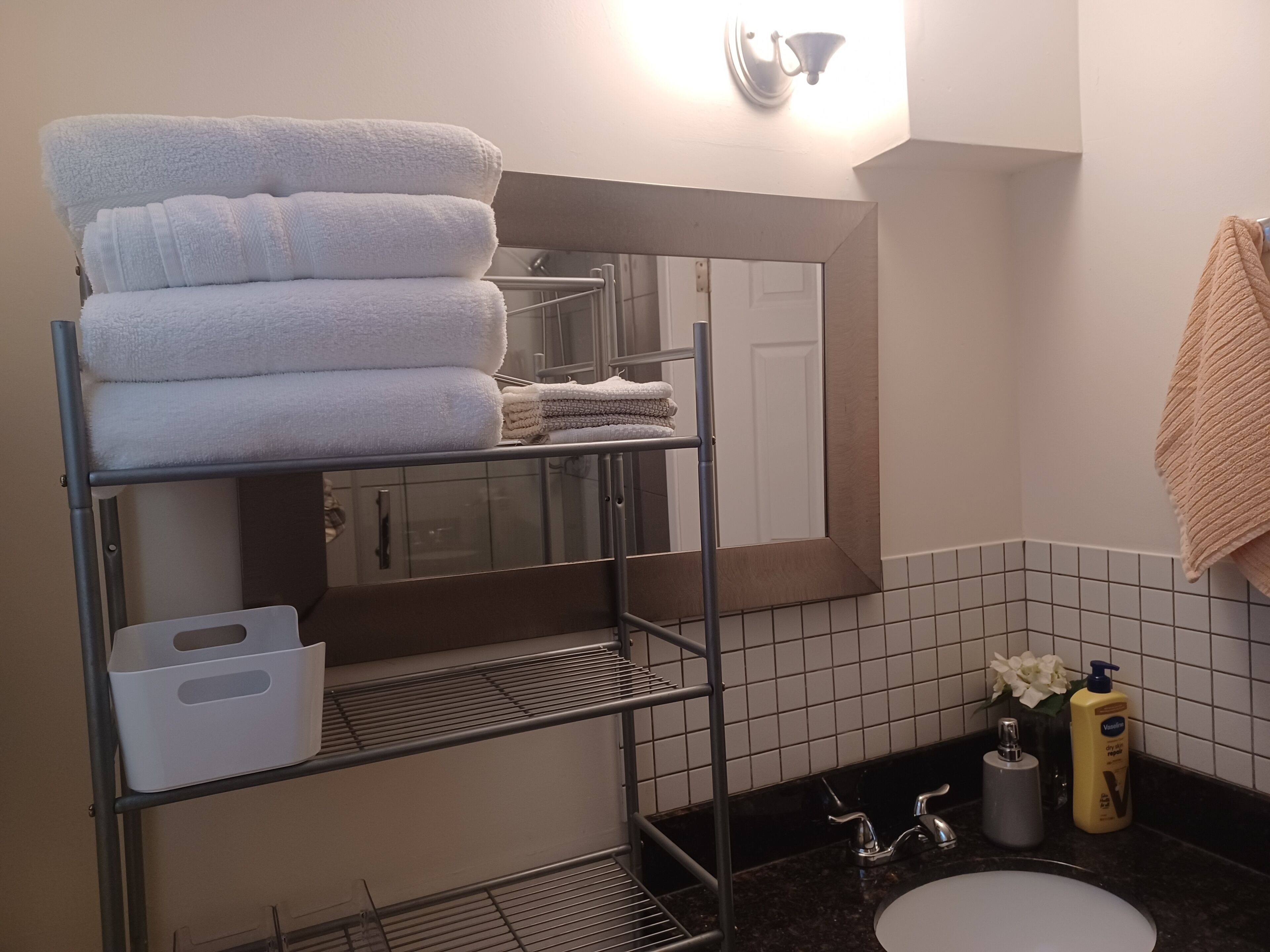 Une salle de bain avec une étagère en métal où sont rangées des serviettes, un miroir, un lavabo et des articles de toilette.
