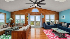 A spacious living room with wooden walls, a ceiling fan, and a large sliding glass door leading to a balcony. - Beachfront Home- Amazing Oceanfront w/ Hot tub!!! Sleeps 8- Free Wifi! -6 beds (Kill Devil Hills)