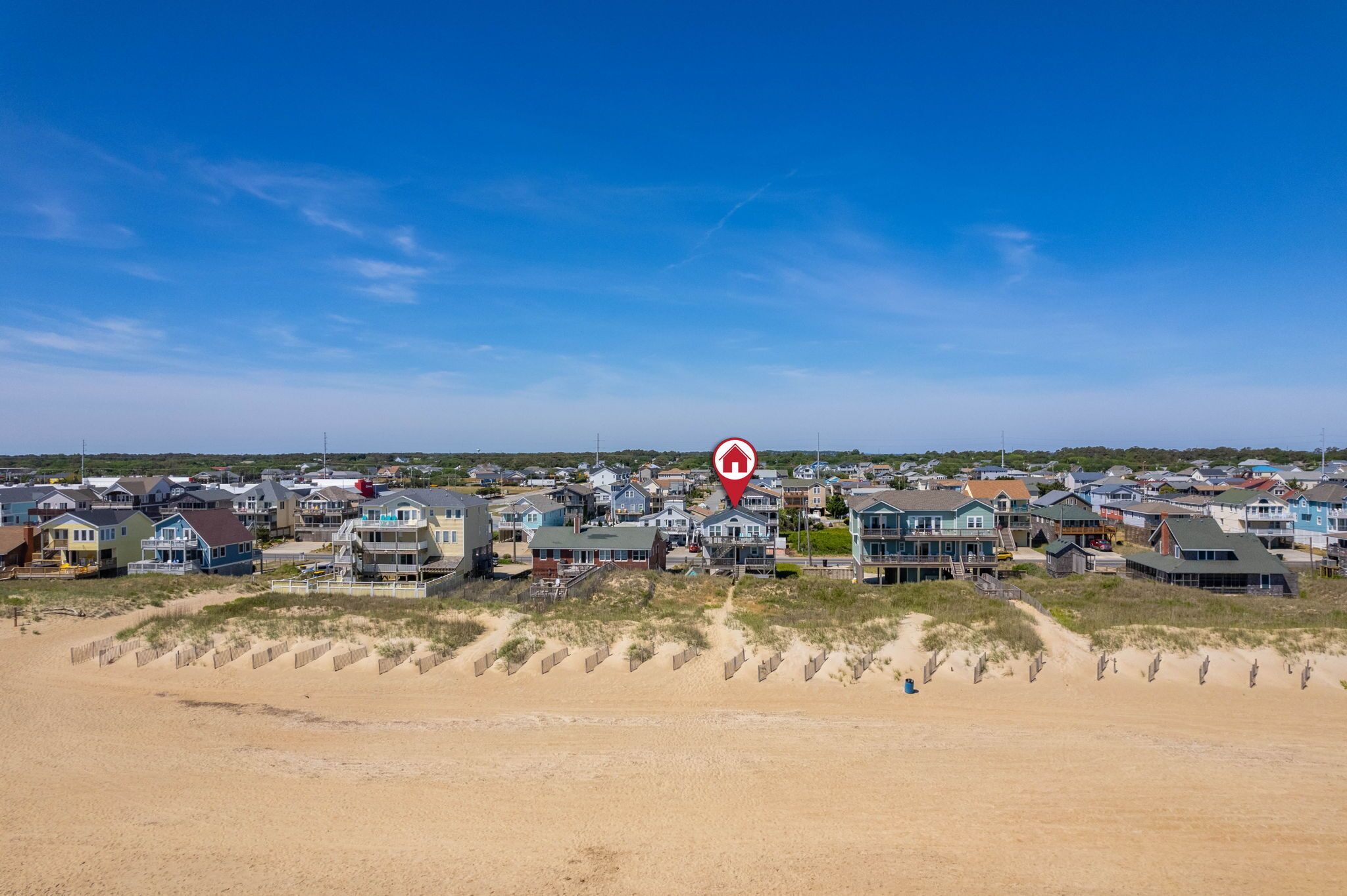 Una zona residencial costera con casas a lo largo de la playa, dunas de arena y un cielo azul despejado.