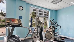 A fitness room with treadmills and elliptical machines, a clock, and a window with a view of palm trees.