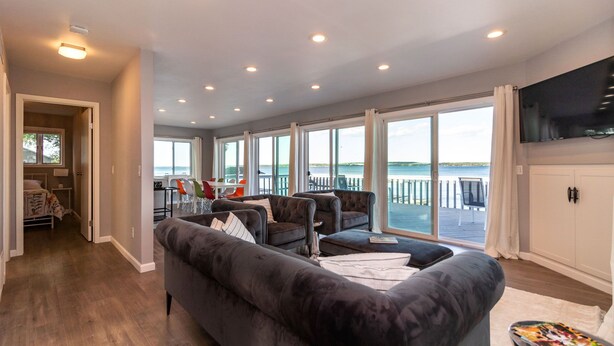 Un salon spacieux avec un canapé gris foncé, un coin repas avec des chaises colorées et de grandes fenêtres offrant une vue sur la mer.