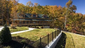A two-story log cabin with a wrap-around porch, surrounded by a well-maintained lawn and a dense forest. - A View to Thrill | Game Room • Hot Tub • Views! (Sevierville)
