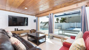 Una sala de estar con techo de madera, un televisor de pantalla plana, un sofá de cuero marrón, un sillón rojo y un ventanal que da a una zona de piscina al aire libre.