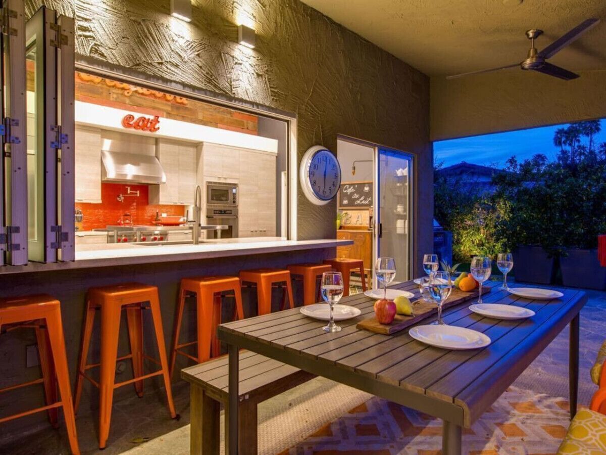 An outdoor dining area with a bar, orange stools, and a table set for two with wine glasses and plates.