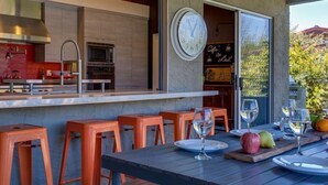 Een terras met houten tafels en oranje stoelen, een klok aan de muur, en uitzicht op de keuken.