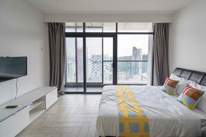 Habitación de hotel moderna con una cama grande, un televisor sobre un mueble bajo y vistas a la ciudad a través de amplios ventanales.