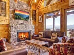 A cozy cabin interior with a stone fireplace, leather furniture, and a large window offering a mountain view.