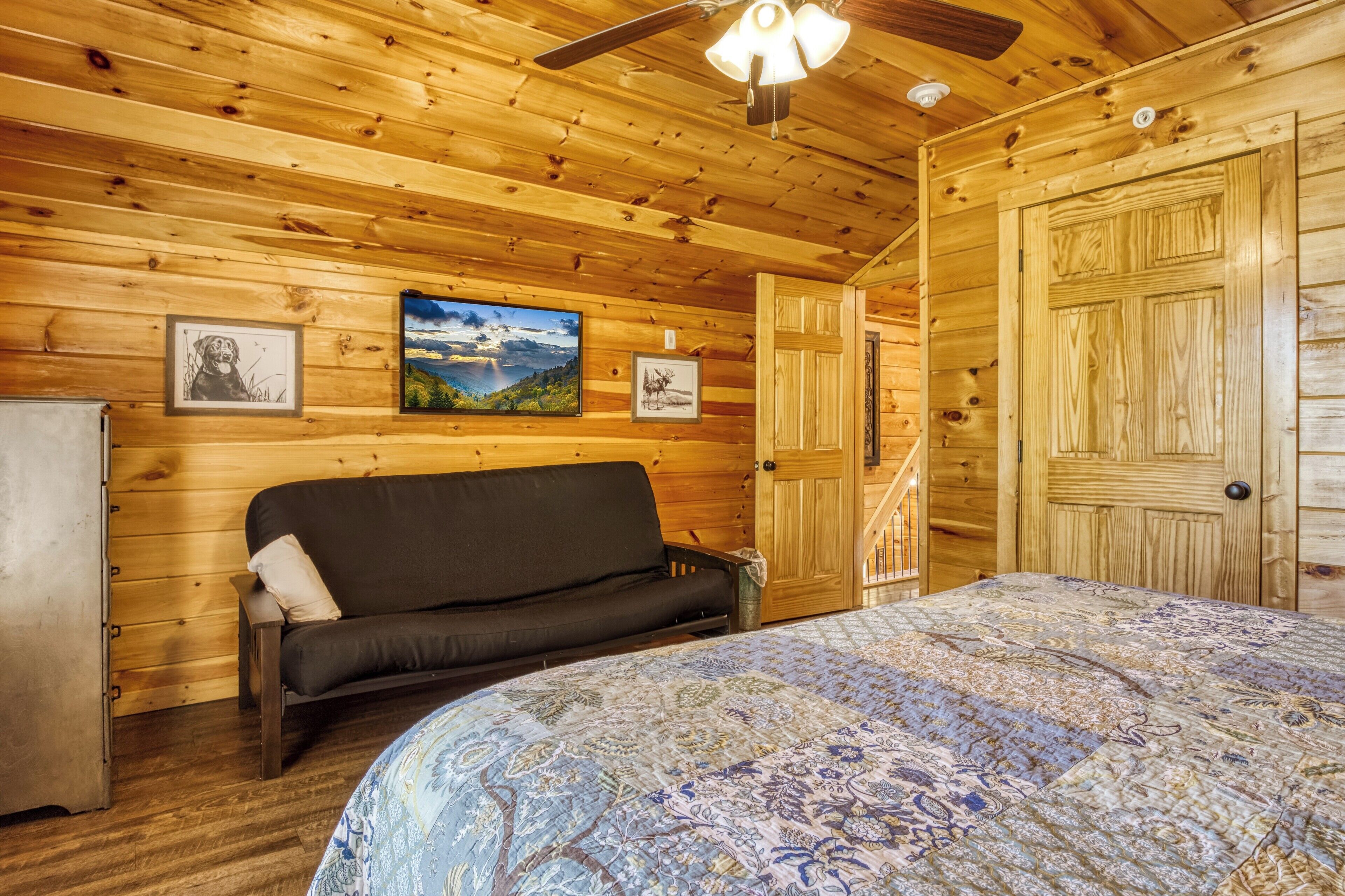 A wooden cabin interior with a bed, a sofa, a ceiling fan, and framed pictures on the walls.