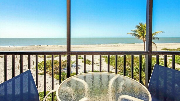 Een balkon met een glazen tafel en stoelen, met uitzicht op een strand met palmbomen en de oceaan.