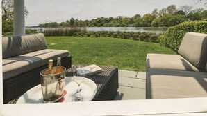 Een terras met tuinmeubilair, een tafel met een glas en fles, en uitzicht op een meer en bomen.