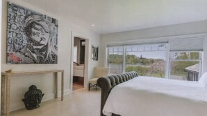 Een slaapkamer met een groot bed, een console met een Boeddhabeeld, een stoel en een groot raam met uitzicht op groen en een brug.