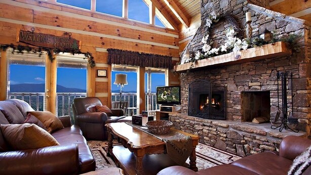 A cozy cabin interior with a stone fireplace, leather furniture, and a large window overlooking a mountainous landscape. - Top-of-Mountain Cabin w/ Views, Hot Tub & Game Room – Near Pigeon Forge (Sevierville)