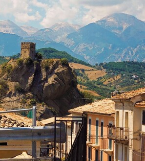 Un paysage montagneux avec un château perché sur un promontoire rocheux, des champs en terrasses et des bâtiments traditionnels dotés de balcons.