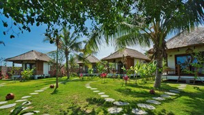 A tropical resort with thatched-roof huts, a well-maintained garden with stepping stones, and palm trees. - Segara Lombok (Selong Belanak)