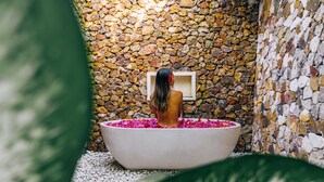 A woman in a bathtub filled with water and rose petals, surrounded by a stone wall. - Segara Lombok (Selong Belanak)