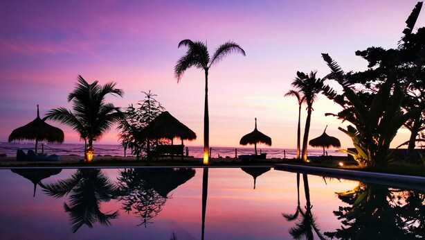 A resort with a pool reflecting palm trees and thatched-roof structures, set against a sunset sky. - Segara Lombok (Selong Belanak)