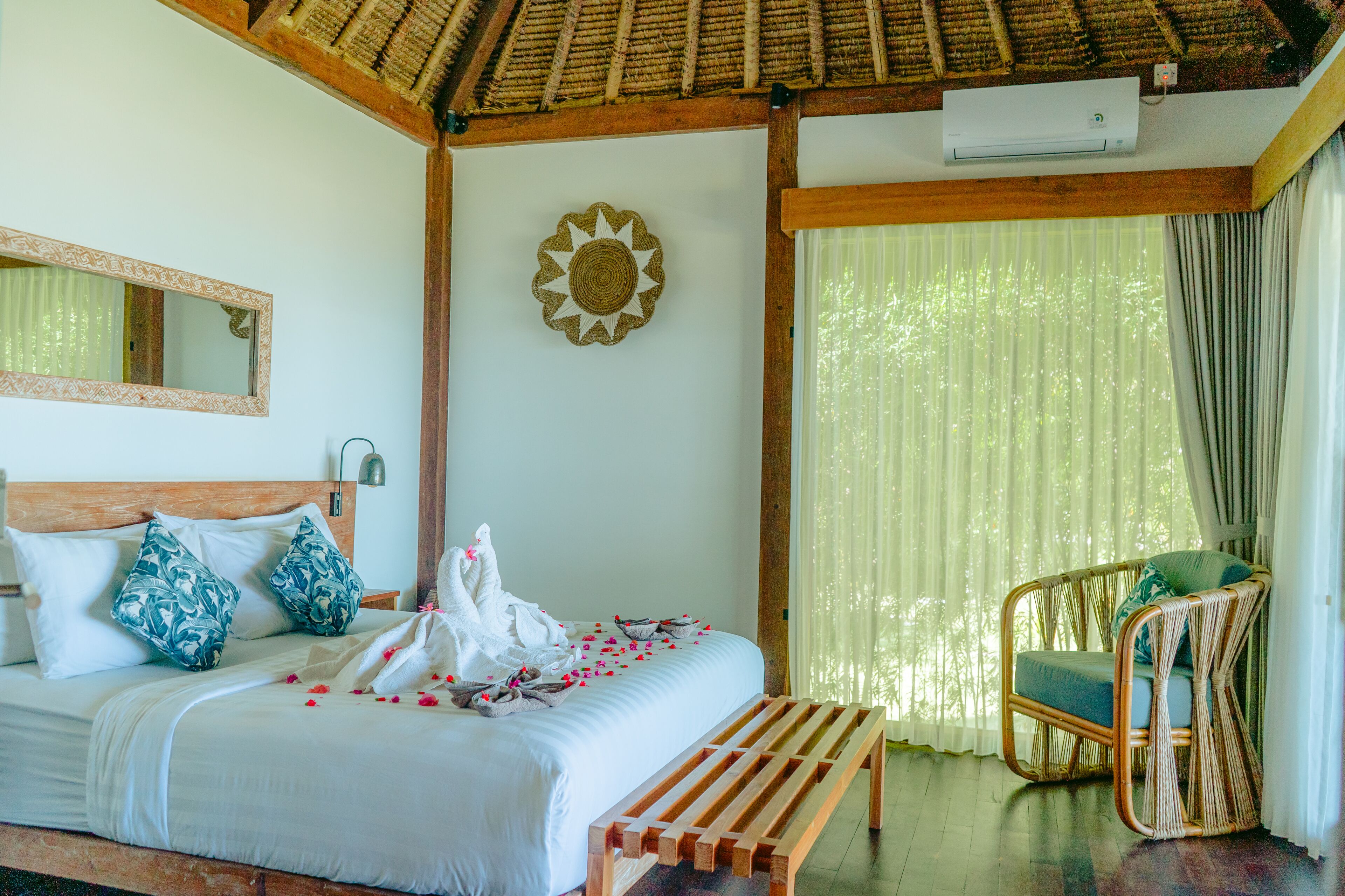 A bedroom with a bed, a wooden bench, a wicker chair, a mirror, and a decorative wall piece.