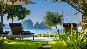 A beachside lounge area with lounge chairs and an umbrella, overlooking the sea with distant rocky formations. - Segara Lombok (Selong Belanak)