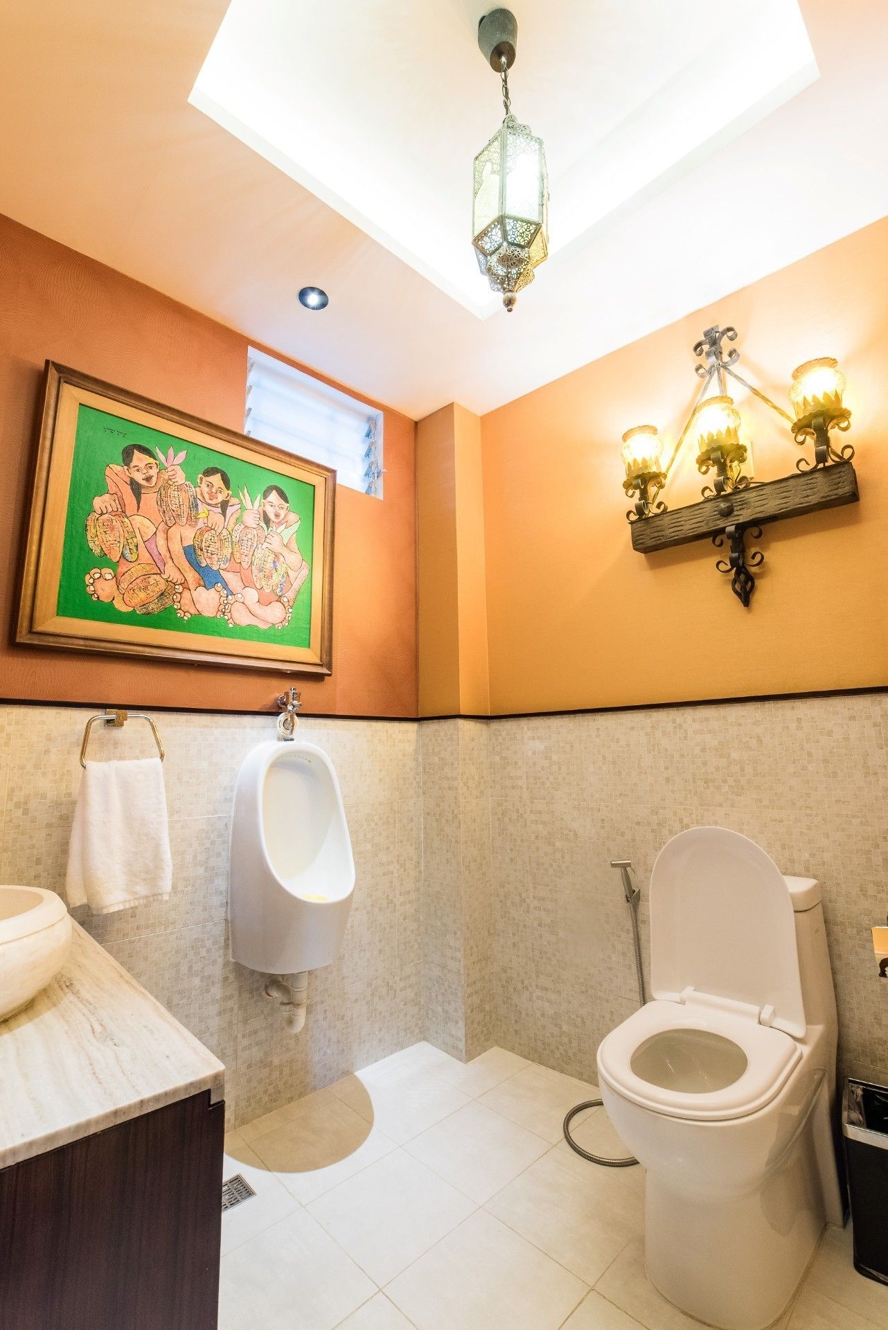Een badkamer met toilet, urinoir en wastafel. Daarboven hangt een decoratieve wandversiering boven de wastafel en een wandlamp boven het toilet.
