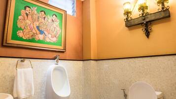Een badkamer met toilet, urinoir en wastafel. Daarboven hangt een decoratieve wandversiering boven de wastafel en een wandlamp boven het toilet.
