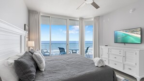 A bedroom with a bed, a TV, a balcony with a view of the sea, and a ceiling fan.