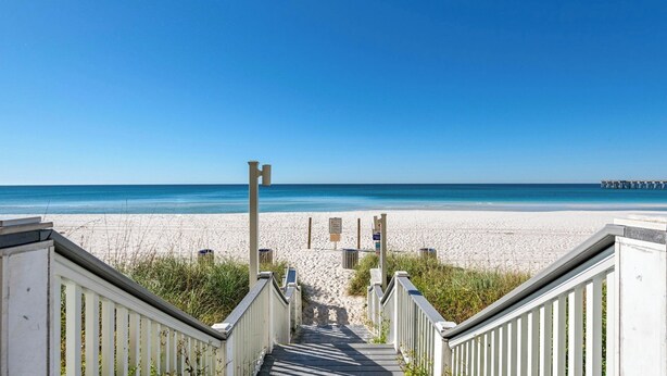 A beach with a wooden staircase leading down to the sand, a pier in the distance, and clear blue skies.