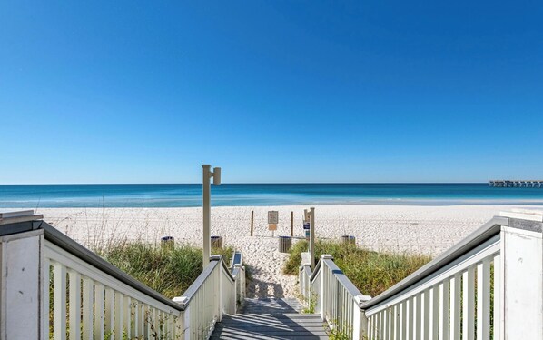 Ein Strand mit einer Holztreppe, die zum Sand führt, einem Pier in der Ferne und blauem Himmel.