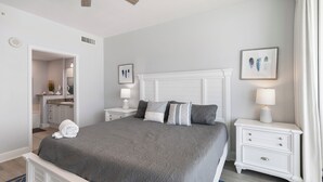 A neatly arranged bedroom with a bed, bedside tables, a nightstand, and a bathroom visible through an open door.