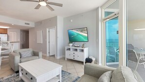 A modern living room with a flat-screen TV, a ceiling fan, a dining area with a table and chairs, and a view of the ocean through a glass door.