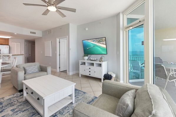 Ein modernes Wohnzimmer mit einem Flachbildfernseher, einem Deckenventilator, einem Essbereich mit Tisch und Stühlen und Meerblick durch eine Glastür.