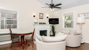 A modern living room with a fireplace, a dining table with chairs, a ceiling fan, and a television mounted on the wall.